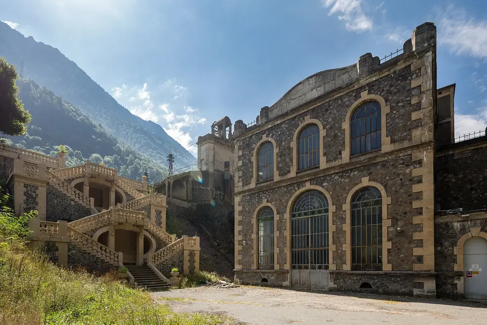 Façade en pierre à grandes baies vitrées d'une ancienne centrale hydroélectrique au pied d'une montagne boisée