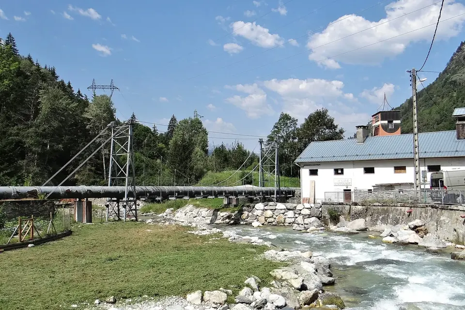 Une conduite forcée franchit un torrent sur une passerelle métallique devant le bâtiment d'une centrale en montagne