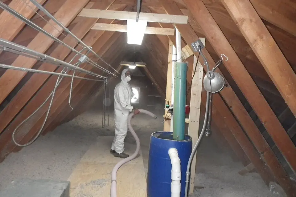 Une personne en combinaison de protection insuffle de l'isolant en vrac dans des combles sous une charpente en bois