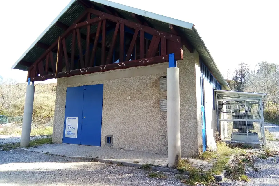 Petit bâtiment technique d'une microcentrale hydroélectrique avec une grande porte bleue, au bord d'un terrain caillouteux