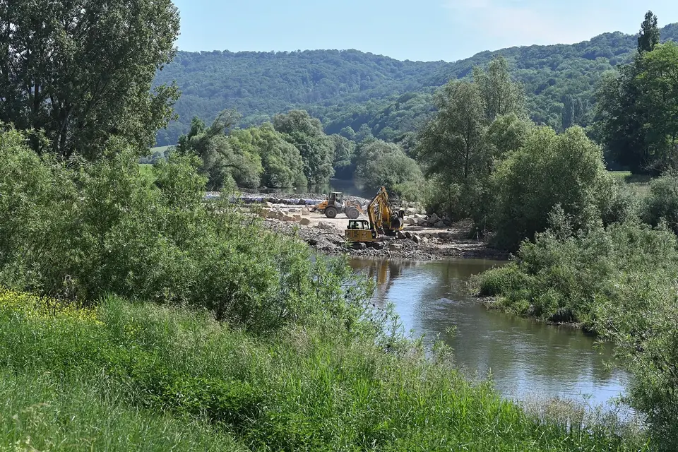Chantier de construction d'une passe à poissons au bord d'une rivière verdoyante, avec une pelleteuse au loin